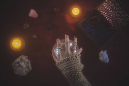 Young woman with a hand on a crystal ball with a mystical background of candles, crystals and stones