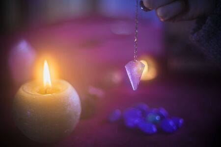 Young woman holding an amethyst pendulum with an esoteric background, candles, stones and crystalsの写真素材