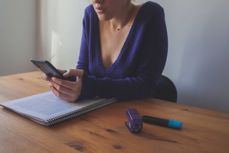 Young woman picking up a mobile phone while studying with highlighters and stapler