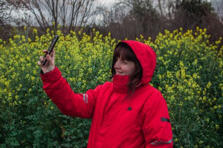 Young woman with red pea coat taking a picture with mobile phone while smiling in the field