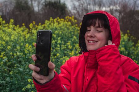 Young woman with red pea coat taking a picture with mobile phone while smiling in the fieldの写真素材