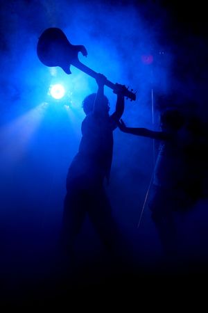 Silhouette of big brother holding guitar up in the air and getting ready to smash it on his little sister, who has her hands out in defense; on stage with fog.の写真素材