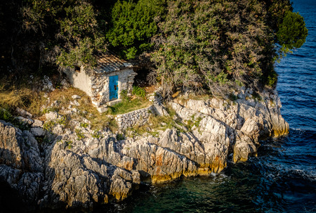 Small old stone lodge on a rocky shore close to a lake, river or sea. Small shed with closed blue door. Next to the dense forest.の写真素材
