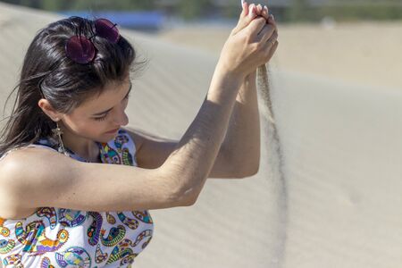 A beautiful young girl sits on the sand in the desert, pours sand from her hands, sand dunes.の写真素材