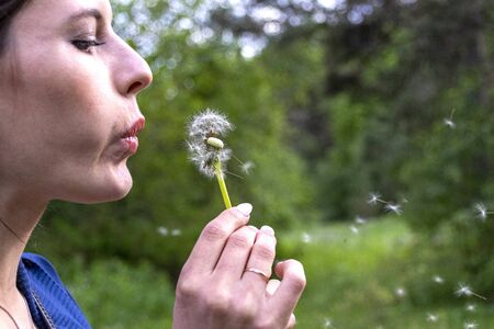 Happy beautiful woman blowing dandelion over sky background, having fun and playing outdoor, teen girl enjoying nature, summer vacation and holidays, young pretty female holding flower, wish concept.の写真素材
