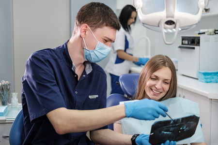 Doctor dentist man in blue robe shows patient to young girl blonde x-ray of teeth in dental office and armchairの写真素材