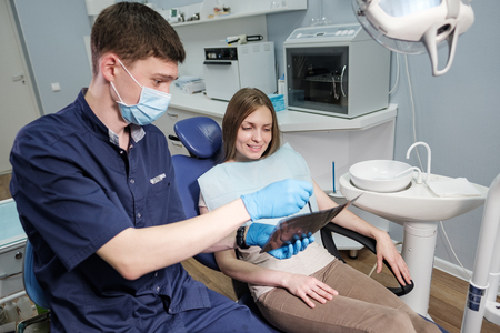 Doctor dentist man in blue robe shows patient to young girl blonde x-ray of teeth in dental office and armchairの写真素材