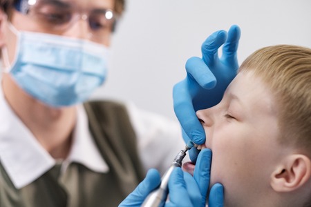 Male dentist examines the teeth of the patient cheerful child with blond hair. Boy smiling in dentists chair with mouth wide open.の写真素材