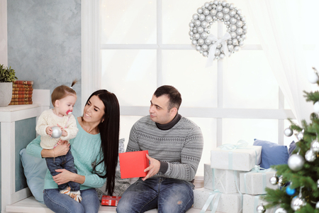 A family of three people chewing on a windowsill with a window on a background of a Christmas tree in sweaters giving presentsの写真素材