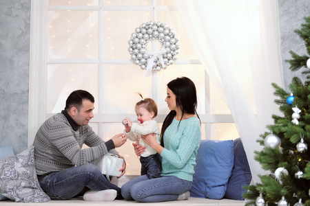 A family of three people chewing on a windowsill with a window on a background of a Christmas tree in sweaters giving presentsの写真素材
