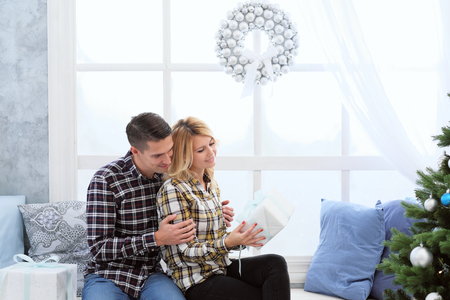 A beautiful young couple in warm checkered shirts sits by the window with New Years decorations and a Christmas tree in blue cold colors and presents gifts in boxes.の写真素材