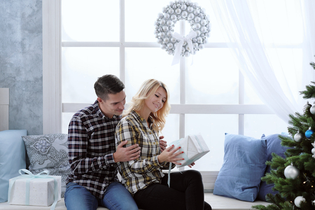 A beautiful young couple in warm checkered shirts sits by the window with New Years decorations and a Christmas tree in blue cold colors and presents gifts in boxes.の写真素材