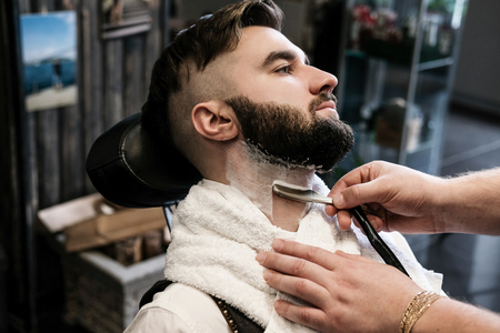 The hairdresser man in glasses cuts and shaves a beard to the young handsome man to the client in a classic suit and a baby girl. Haircut in the salon, barbershopの写真素材
