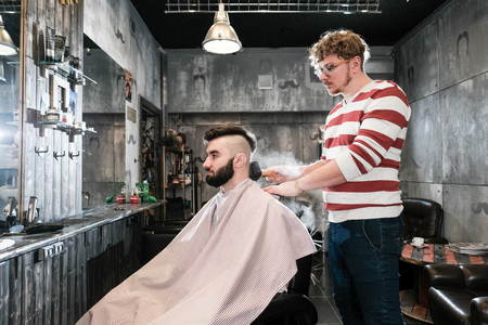 The hairdresser man in glasses cuts and shaves a beard to the young handsome man to the client in a classic suit and a baby girl. Haircut in the salon, barbershopの写真素材