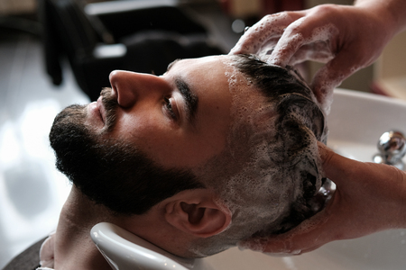 The hairdresser man in glasses cuts and shaves a beard to the young handsome man to the client in a classic suit and a baby girl. Haircut in the salon, barbershopの写真素材