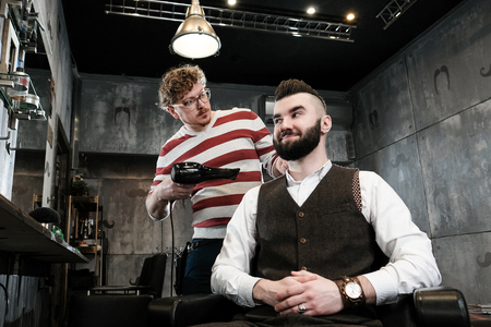 The hairdresser man in glasses cuts and shaves a beard to the young handsome man to the client in a classic suit and a baby girl. Haircut in the salon, barbershopの写真素材