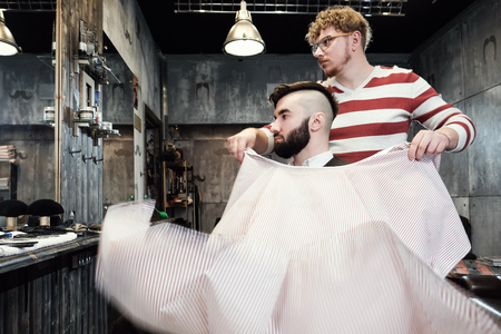 The hairdresser man in glasses cuts and shaves a beard to the young handsome man to the client in a classic suit and a baby girl. Haircut in the salon, barbershopの写真素材