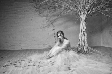 A young blonde bride in white lace designer wedding dress on a background of white brick wall and...