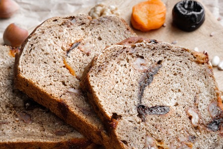 Rye bread with nuts, dried fruits, apricots and prunes, dessert. The whole loaf and the cut pieces of bread lie on brown paper on a table made of ebony. Nearby lie walnuts and hazelnuts, dried apricots and prunes. Warm key, many objects.の写真素材
