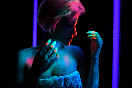 Young beautiful girl model with short haircut in the studio on a black background. Neon paints, makeup, body painting, colored hair coloring, creative color, poses in the light of soffits. Hands, face, blue, green, orange.の写真素材