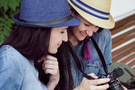 Two young beautiful girls in jeans dresses and hats sit on a bench in the park on a background of green plant walls, and are photographed. They look at the photos on the screen, choose frames, discuss and laugh.の写真素材