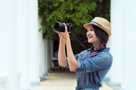 Young beautiful girl photographer taking a photograph of a close-up. Against the background of a green wall of plants. A park. Hat and denim dress. Sale of phototechnicsの写真素材