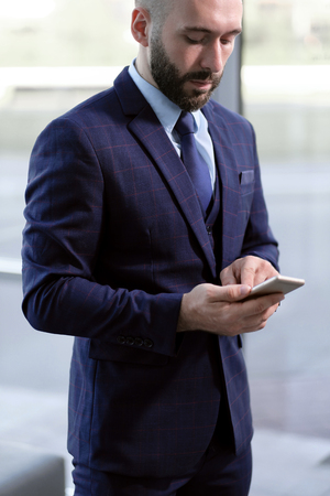 A handsome male businessman uses a smartphone on the background of the entrance to an office building. Blue business suit, daylight.の写真素材