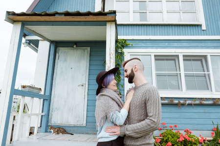 Portrait of young woman and a man, a family smiling beautiful woman with blue hair sitting on porch of country blue house, green loach plant and vase with sunflowers, yellow pumpkins. A sweater, scarf and hat.の写真素材