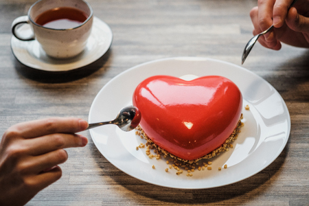 Red cake in the shape of a heart is on the table. Two hands with spoons, male and female, stretch into a pie. Romantic date, Valentine's Day.の写真素材