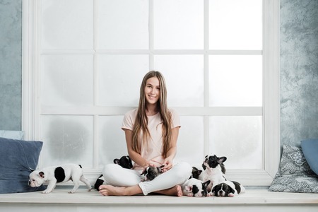 A beautiful young girl sits on the windowsill of a white window and holds a lot of small puppy of a French bulldog dog.の写真素材