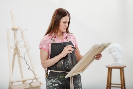 Young woman artist draws a pencil on canvas. White studio, pink shirt and apron. Drawing and painting lessons, professional artist.の写真素材