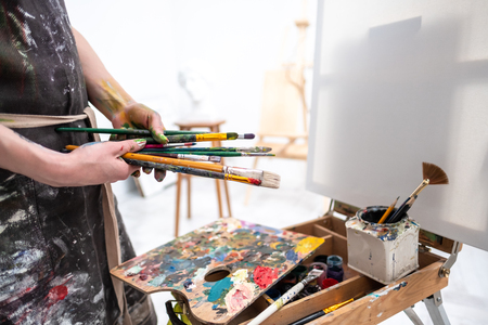 Young beautiful girl painter in a white studio draws on an easel on canvas. Draws a plaster head. Pink checked shirt and black apron.の写真素材
