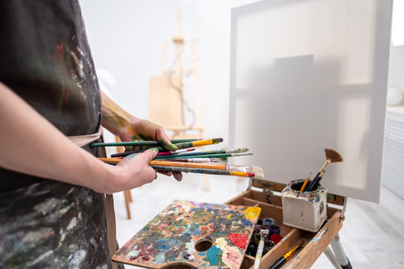Young beautiful girl painter in a white studio draws on an easel on canvas. Draws a plaster head. Pink checked shirt and black apron.の写真素材