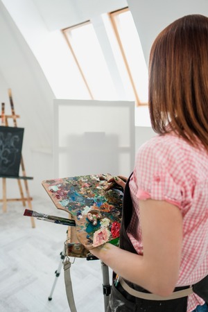 Young beautiful girl painter in a white studio draws on an easel on canvas. Draws a plaster head. Pink checked shirt and black apron.の写真素材