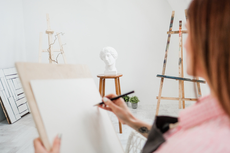 Young woman artist draws a pencil on canvas. White studio, pink shirt and apron. Drawing and painting lessons, professional artist.の写真素材