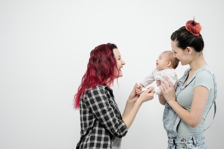 Two young women with dyed red hair and in casual clothes with a baby on a white background. Same-sex marriage and adoption, homosexual lesbian couple.の写真素材