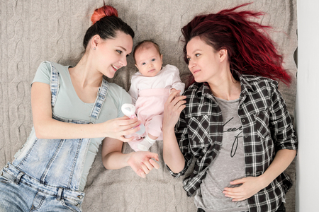 Two young women in casual clothes and with pink hair, a lesbian homosexual couple, lying on a rug with a child. Same-sex marriage, adoption.の写真素材