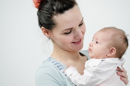 Young woman mother in denim overalls holds a baby child in her arms. White background in the studio. Happy family, baby smiling.の写真素材