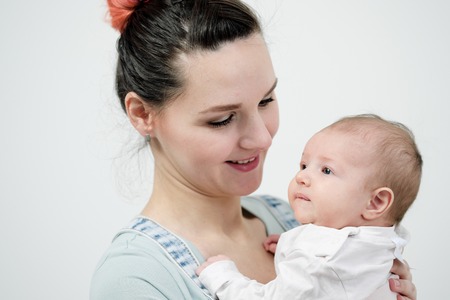 Young woman mother in denim overalls holds a baby child in her arms. White background in the studio. Happy family, baby smiling.の写真素材