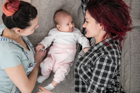 Two young women in casual clothes and with pink hair, a lesbian homosexual couple, lying on a rug with a child. Same-sex marriage, adoption.の写真素材