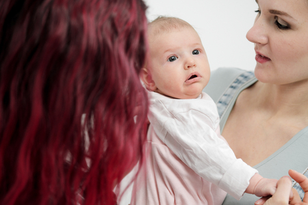 Two young women with dyed red hair and in casual clothes with a baby on a white background. Same-sex marriage and adoption, homosexual lesbian couple.の写真素材