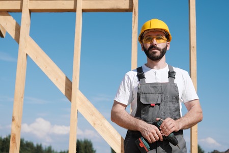 Portrait of a male builder in overalls and a yellow helmet on the roof of a frame house with a...