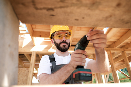 Portrait of a male builder in overalls and a yellow helmet on the roof of a frame house. Screws with a screwdriver.の写真素材