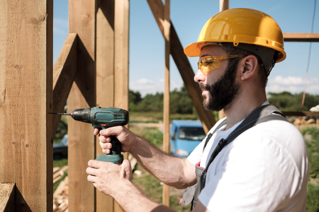 Portrait of a male builder in overalls and a yellow helmet on the roof of a frame house. Screws with a screwdriver.の写真素材