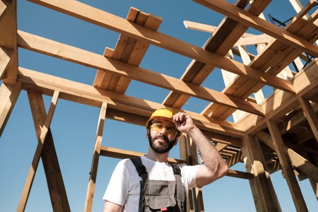 The man is a builder on the background of the roof of a frame house, in a yellow helmet and gray overalls.の写真素材