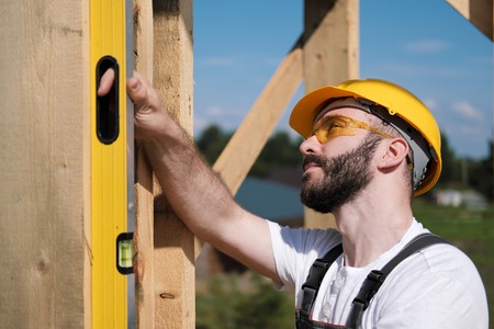 Portrait of a male builder in overalls and a yellow helmet on the roof of a frame house. Produces measurements with the help of a building level.の写真素材