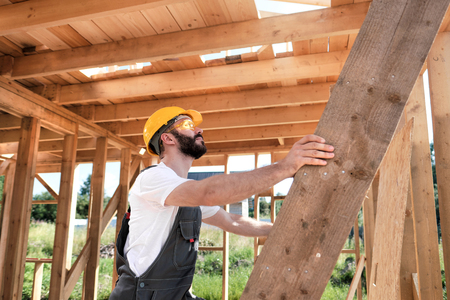 Portrait of a male builder in overalls and a yellow helmet on the roof of a frame house. He climbs the stairs.の写真素材