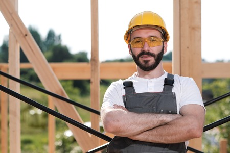 The man is a builder on the background of the roof of a frame house, in a yellow helmet and gray overalls.の写真素材