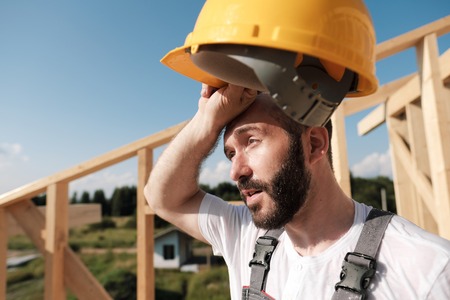 The man is a builder on the background of the roof of a frame house, in a yellow helmet and gray overalls.の写真素材