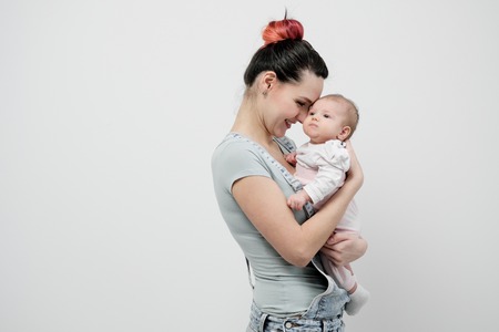 Young woman mother in denim overalls holds a baby child in her arms. White background in the studio. Happy family, baby smiling.の写真素材